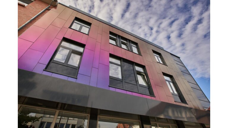 Fassade mit irisierenden Paneelen in Violett- und Pinktönen sowie großen Fenstern, vor einem teils bewölkten Himmel.